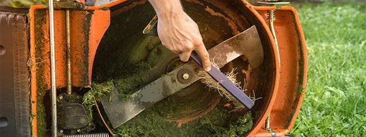 Hand cleaning grass clippings from the blade of an orange lawn mower on green grass
